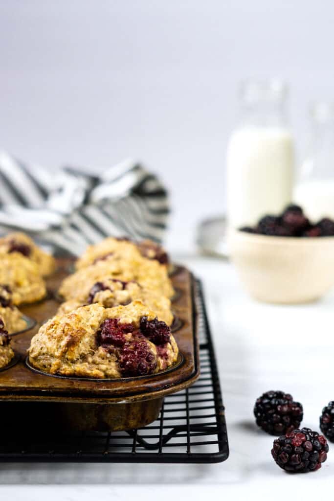 Blackberry oatmeal muffins cooling in a pan on a cooling rack beside a bowl of fresh blackberries and a small glass jug of milk.