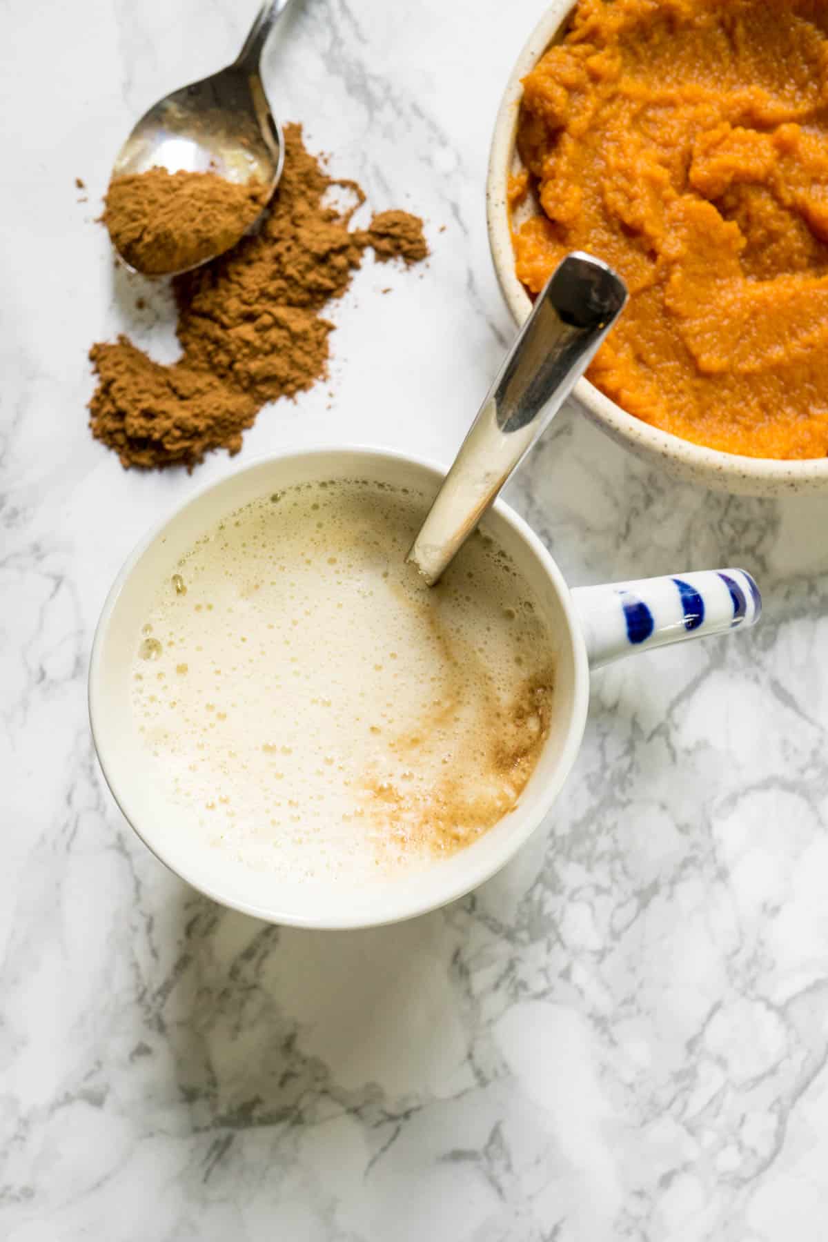 A mug of homemade pumpkin spice latte with a spoonful of spices and a bowl of pumpkin puree.