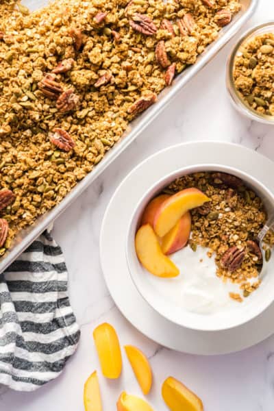 a sheet pan of pumpkin maple pecan granola beside a bowl with yogurt, granola, and peaches