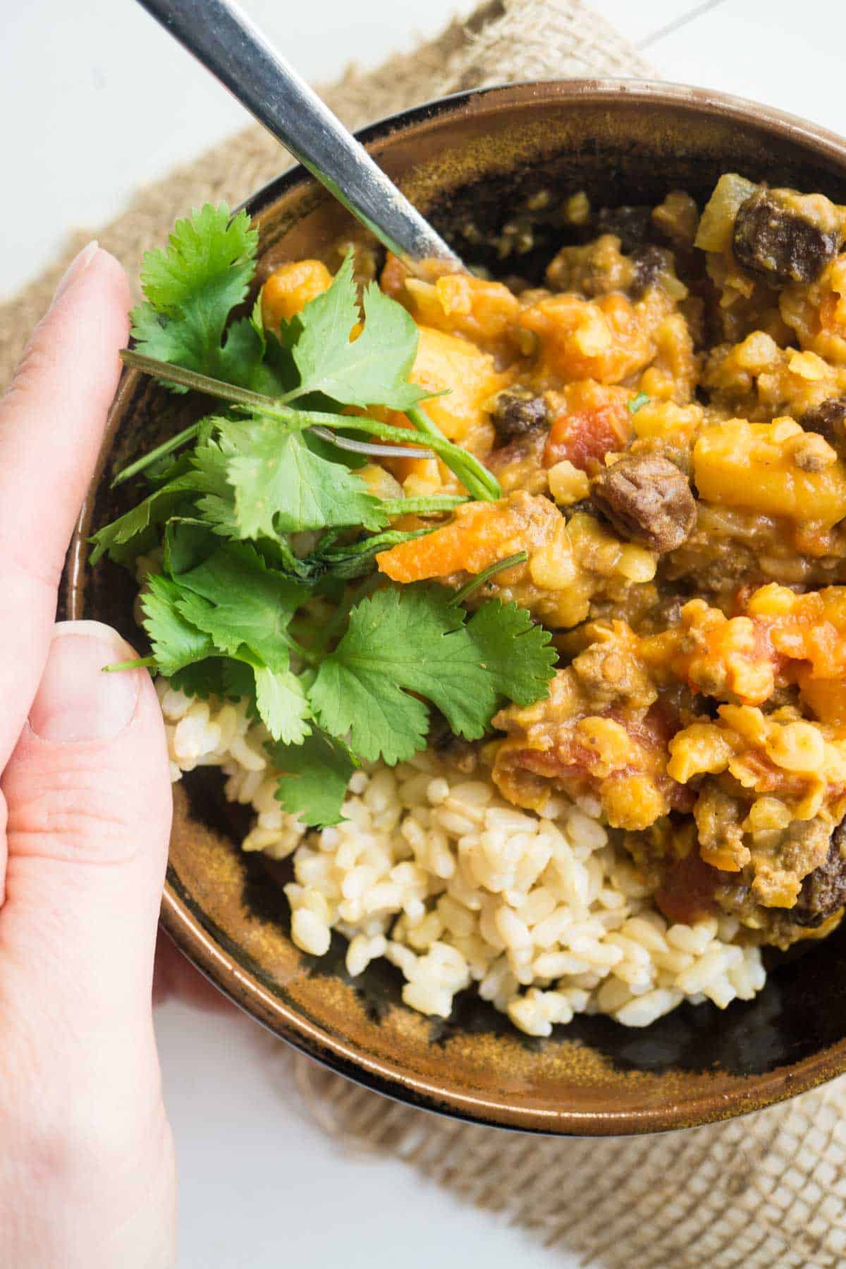 Moroccan Beef and Lentil Bowl with parsley garnish and rice. A hand holding the side of the bowl