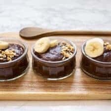 3 small bowls of chocolate pudding on a wooden board topped with sliced bananas and chopped peanuts