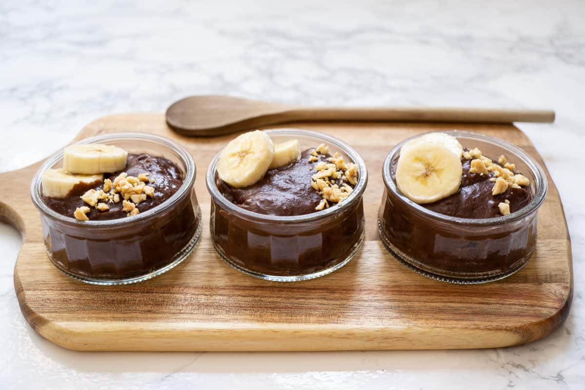 3 small bowls of chocolate pudding on a wooden board topped with sliced bananas and chopped peanuts