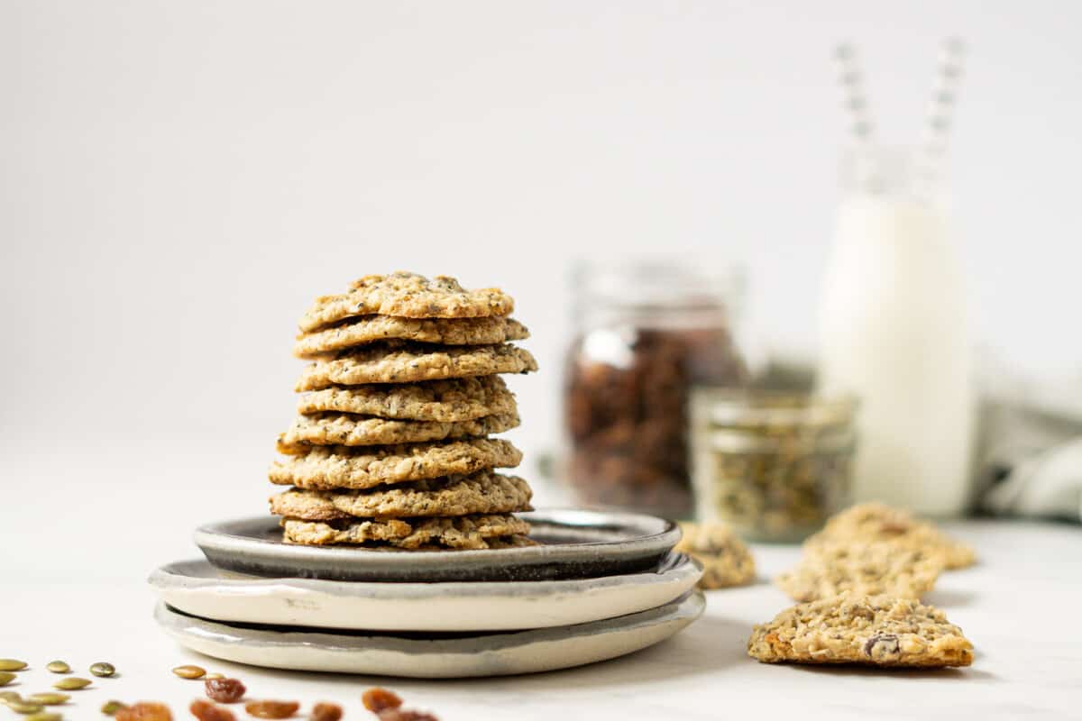 a plate of stacked trail mix cookies in front of a glass of milk and jars of raisins and pumpkin seeds