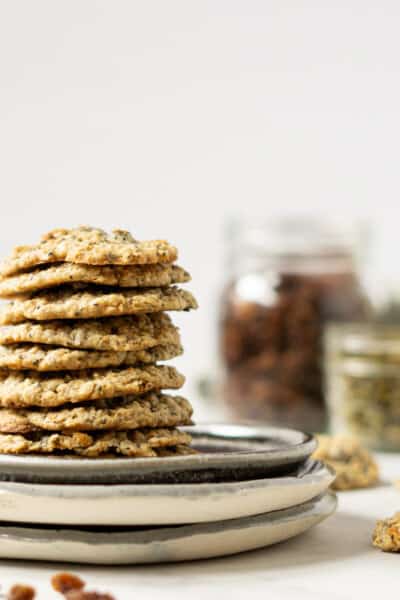 a plate of stacked trail mix cookies in front of a glass of milk and jars of raisins and pumpkin seeds