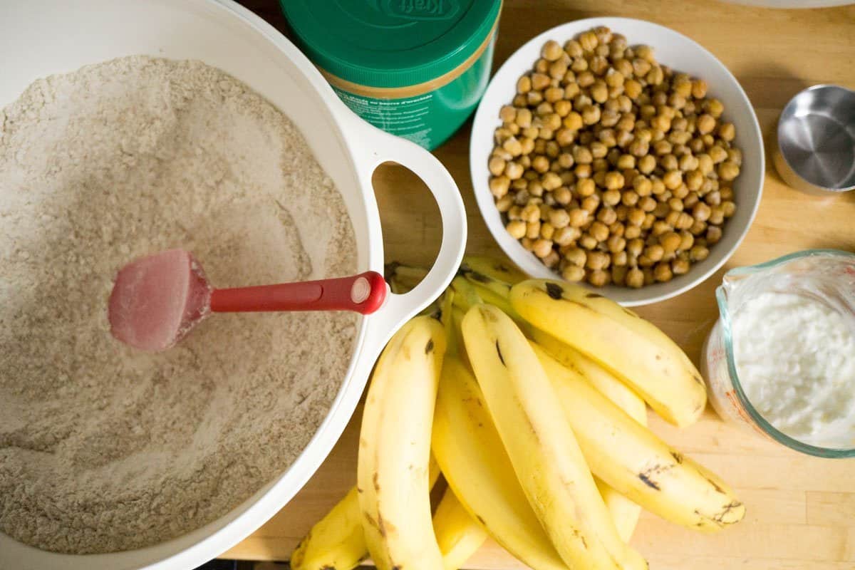 Ingredients set out on a counter for big batch protein muffins: peanut butter, chickpeas, bananas, and flour