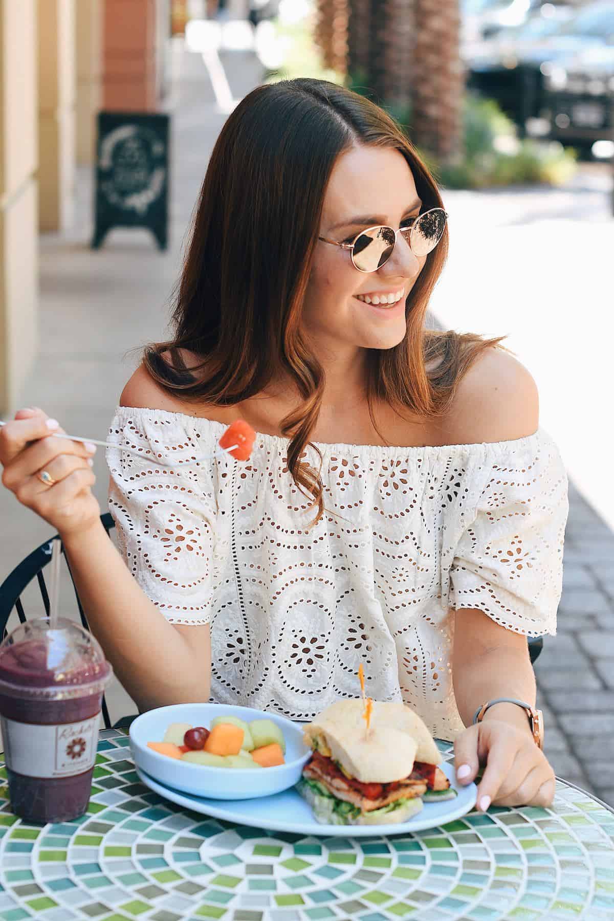 woman wearing sunglasses sitting at an outdoor cafe eating a fruit salad