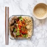 Ginger beef, veggies, brown rice in a meal prep container with toasted sesame seeds and chopsticks