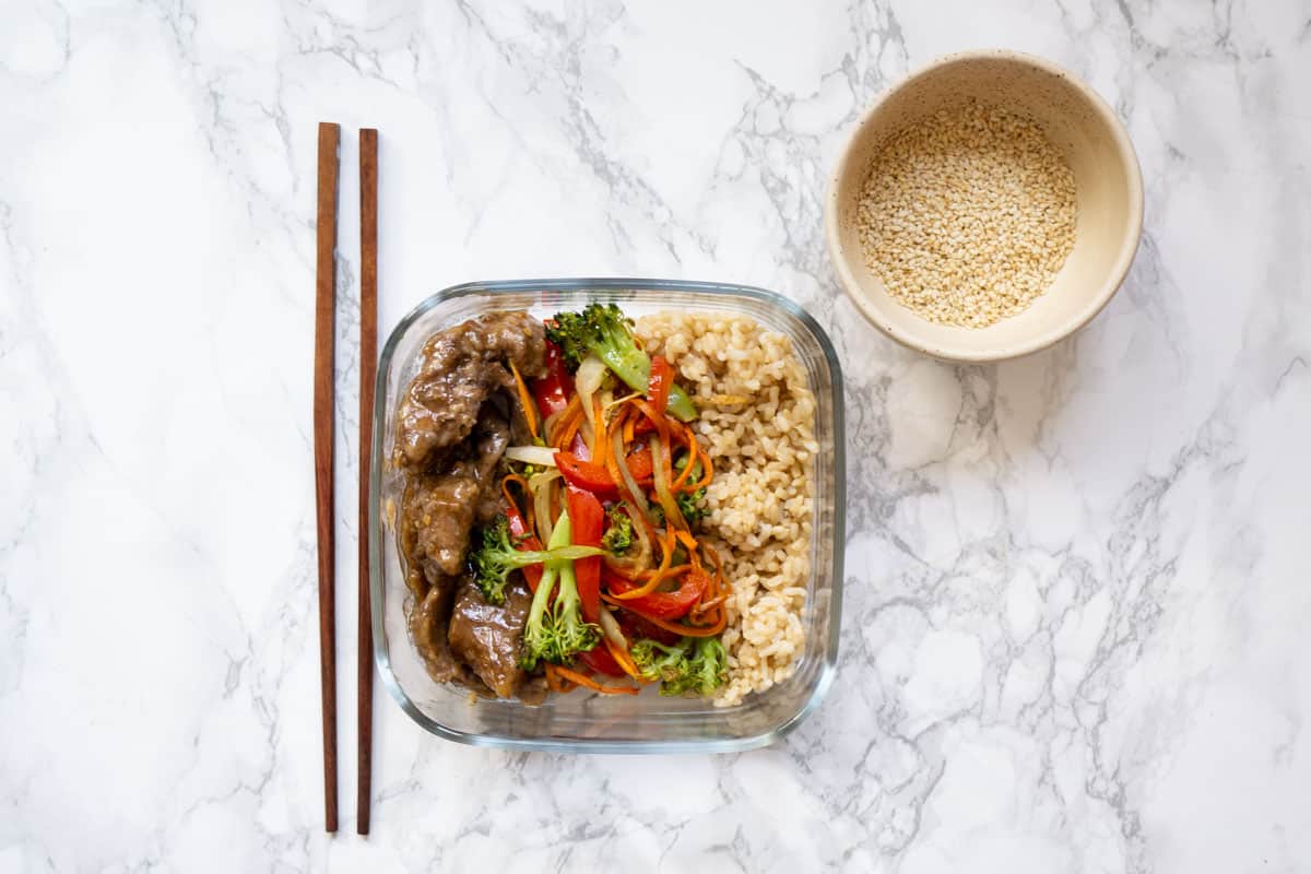 Ginger beef, veggies, brown rice in a meal prep container with toasted sesame seeds and chopsticks