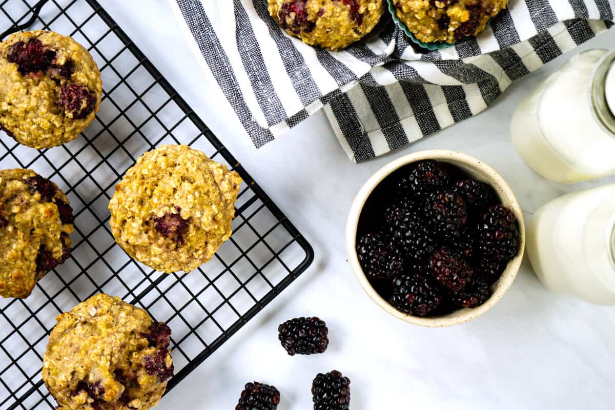 overhead view of blackberry oatmeal muffins cooling on a rack beside a bowl of fresh blackberries and two glass jars of milk