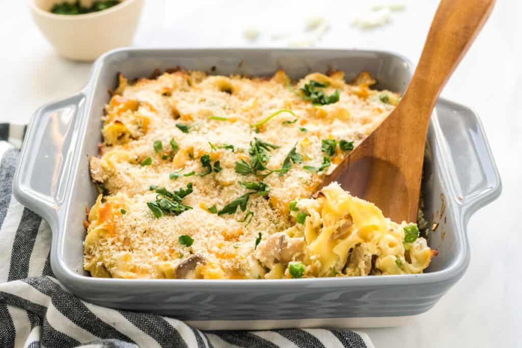 A close up view of a serving spoon scooping into a baking dish filled with creamy tuna noodle casserole topped with panko crumbs.