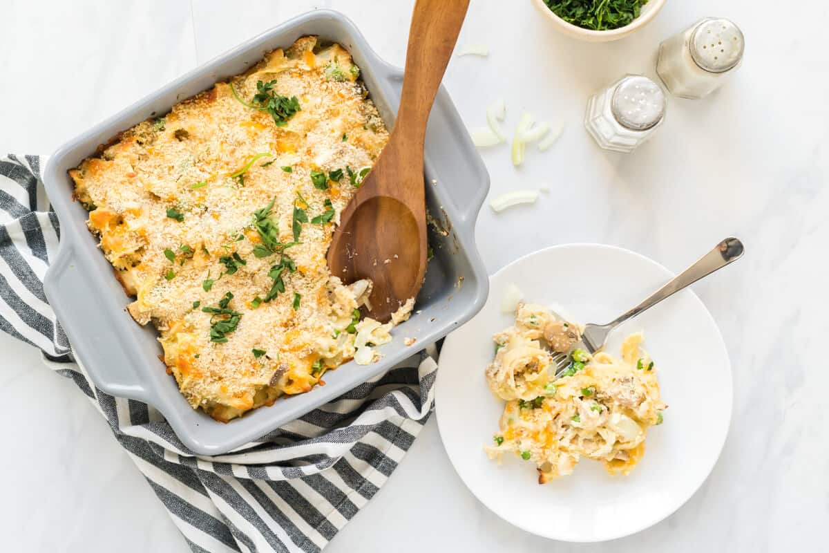 Baking dish with creamy tuna noodle casserole with a serving spoon that has scooped out a plateful, which is right beside the baking dish.