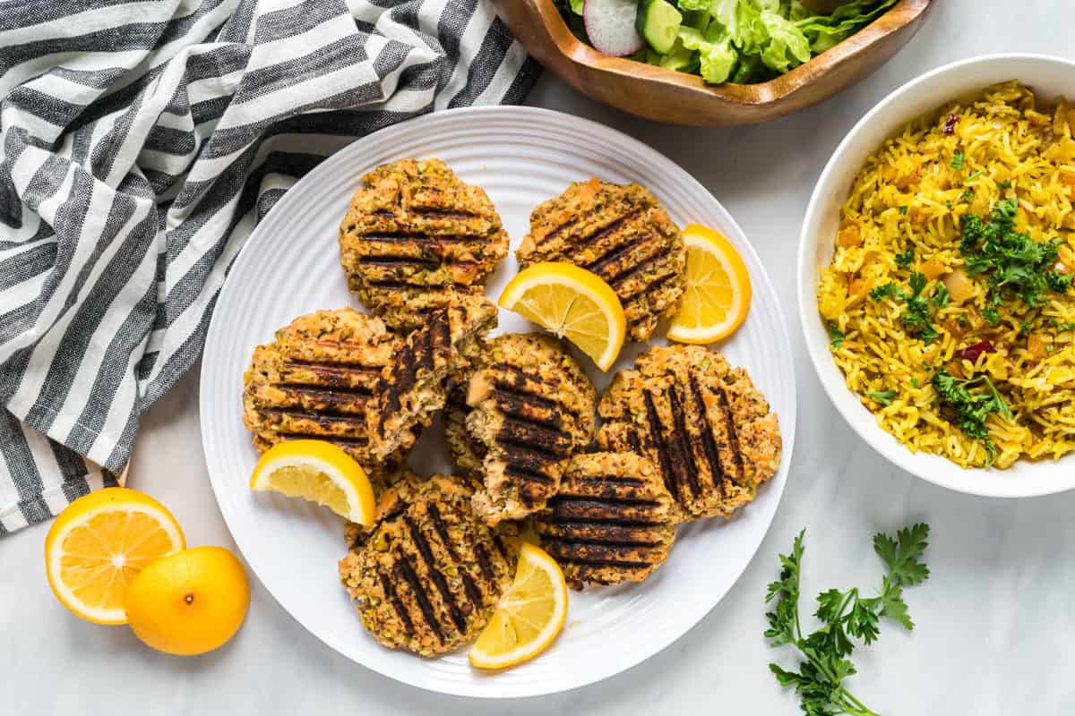 A plate of pistachio salmon cakes with a salad and a bowl of apricot rice to the side.