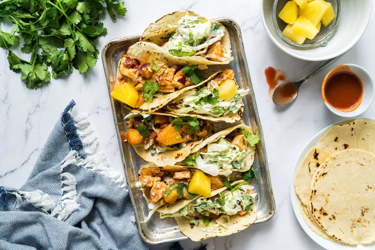 Flatlay view of a sheetpan of pineapple chicken tacos with cilantro slaw with a bowl of pineapple, stack of corn tortillas, and fresh cilantro.