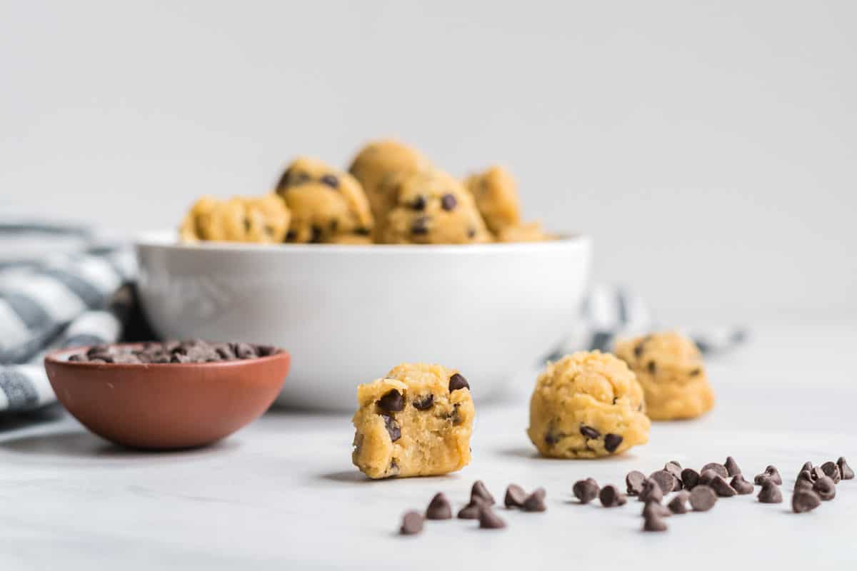 A bowl of edible chocolate chip cookie dough bites with a few beside. One has a bit taken out to show the texture.