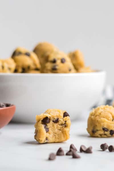 A bowl of edible chocolate chip cookie dough bites with a few beside. One has a bit taken out to show the texture.