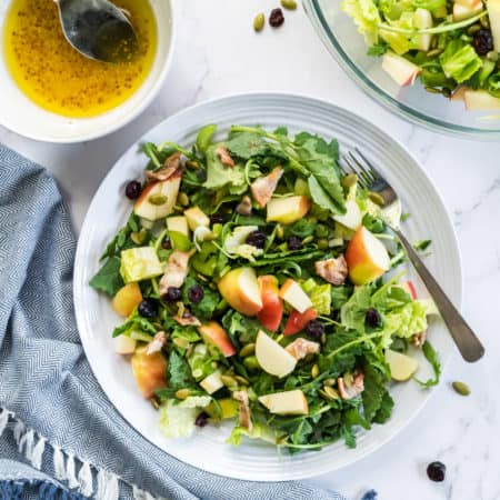 flatlay view of a plat of bacon apple arugula salad with cranberries, pumpkin seeds and a bowl of lemon vinaigrette on the side