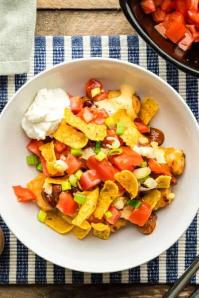 A bowl of Disney Totchos surrounded by toppings: chili, diced tomatoes, sliced green onions, and corn chips.