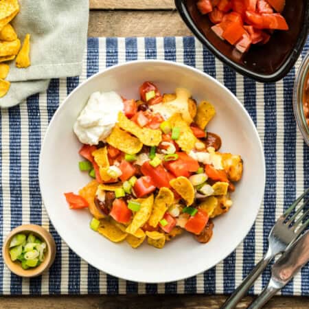 A bowl of Disney Totchos surrounded by toppings: chili, diced tomatoes, sliced green onions, and corn chips.